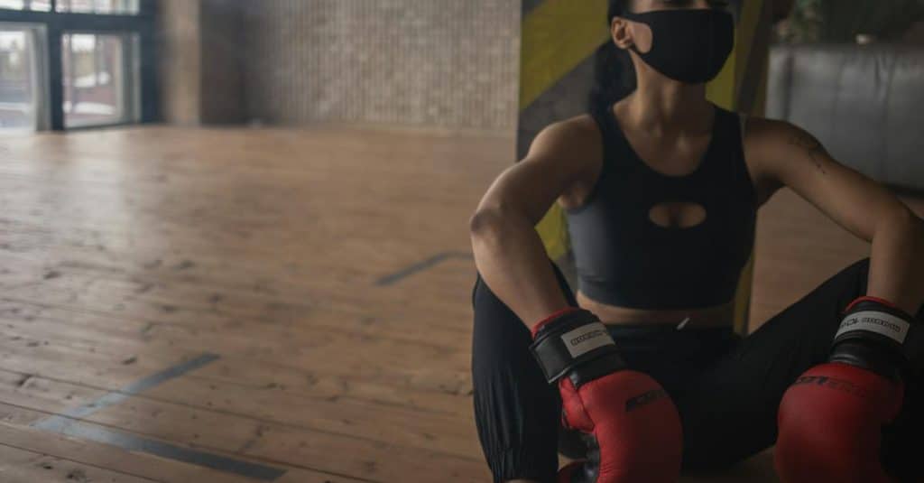 african-american-female-boxer-in-protective-mask-resting-on-floor-after-training-in-fitness-center